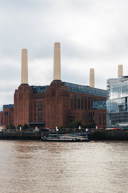 A low-angle view of the Battersea Power Station in London, featuring its historic red-brick facade with tall, narrow windows and white chimney stacks extending into a partly cloudy sky. Surrounding th