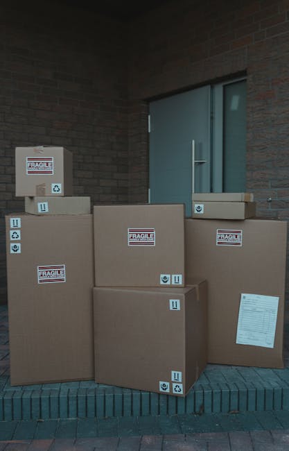 A group of five large cardboard boxes, some labeled with 'Fragile' stickers and handling symbols, are positioned on a brick doorstep next to a brick building with a closed entry door. The boxes vary i