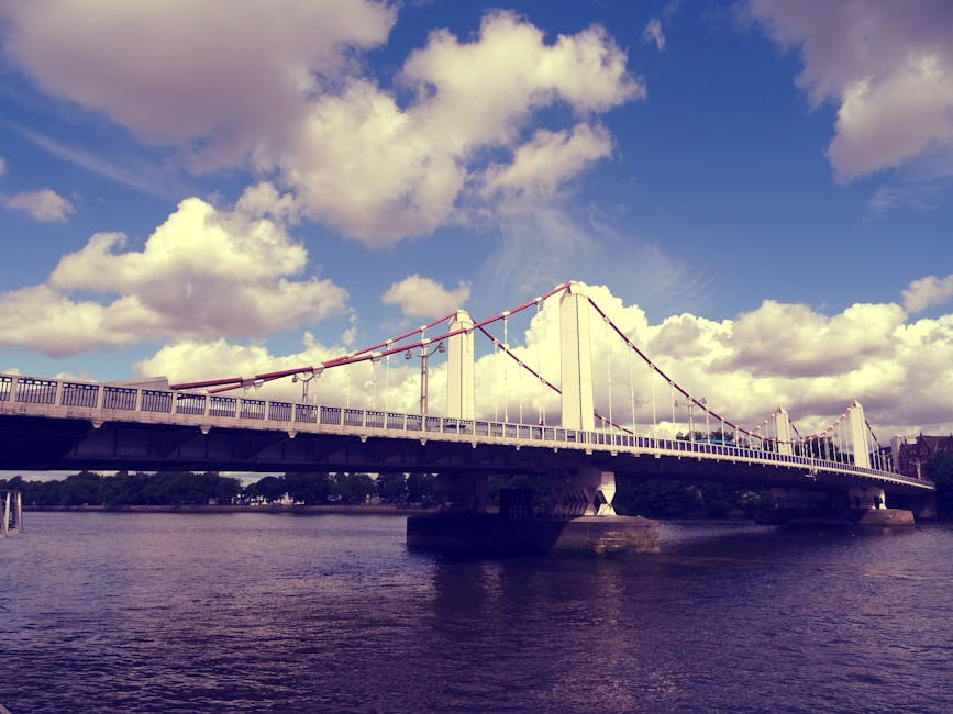 A large suspension bridge spanning a wide river under a partly cloudy sky, with white towers and red suspension cables. The bridge has a roadway with a guardrail and is supported by several concrete p